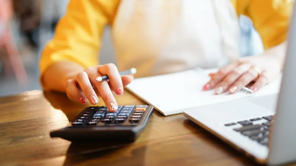 Woman using calculator and notebook to calculate prices for her online boutique.