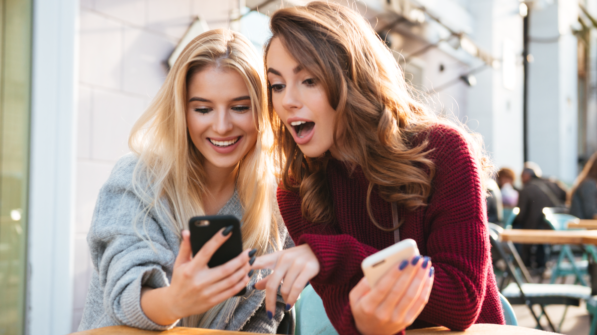 Two friends excitedly share their phone screens to introduce their favorite brands through word-of-mouth marketing.
